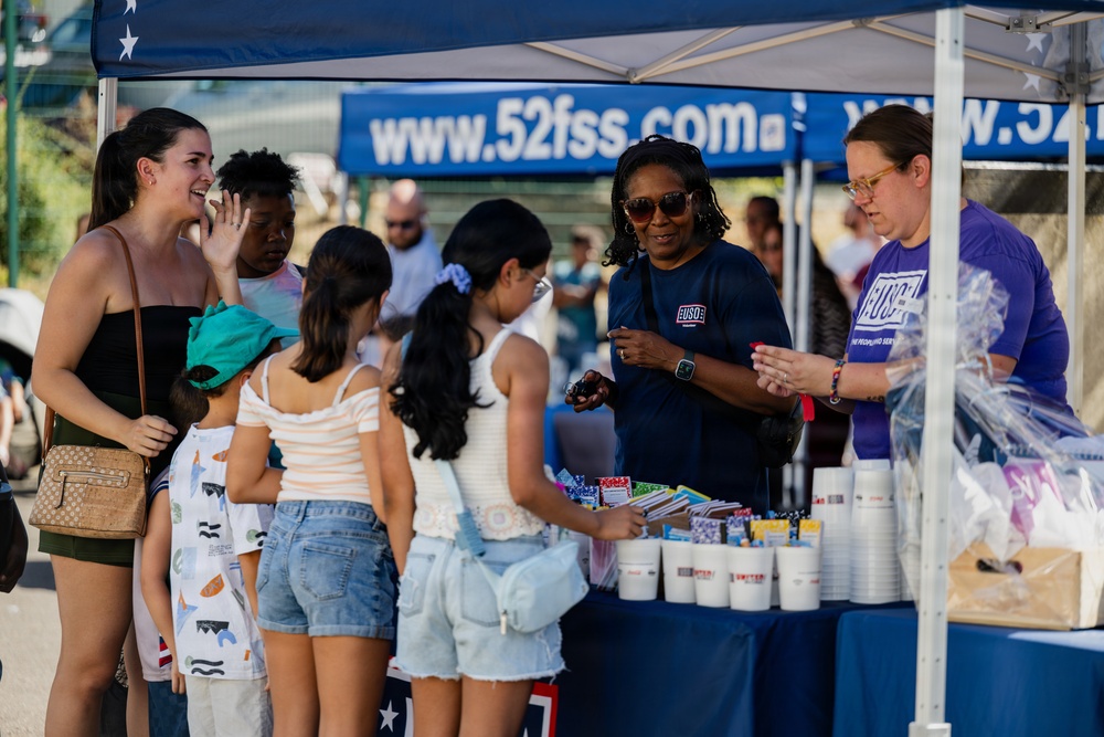 A USO booth at what appears to be an outdoor community or military base event, operating under a blue canopy tent branded with 'www.52fss.com'. Two USO volunteers in branded t-shirts are distributing items to a group of visitors including adults and children. The atmosphere is casual and festive, suggesting a family-friendly public gathering.