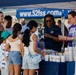 A USO booth at what appears to be an outdoor community or military base event, operating under a blue canopy tent branded with 'www.52fss.com'. Two USO volunteers in branded t-shirts are distributing items to a group of visitors including adults and children. The atmosphere is casual and festive, suggesting a family-friendly public gathering.