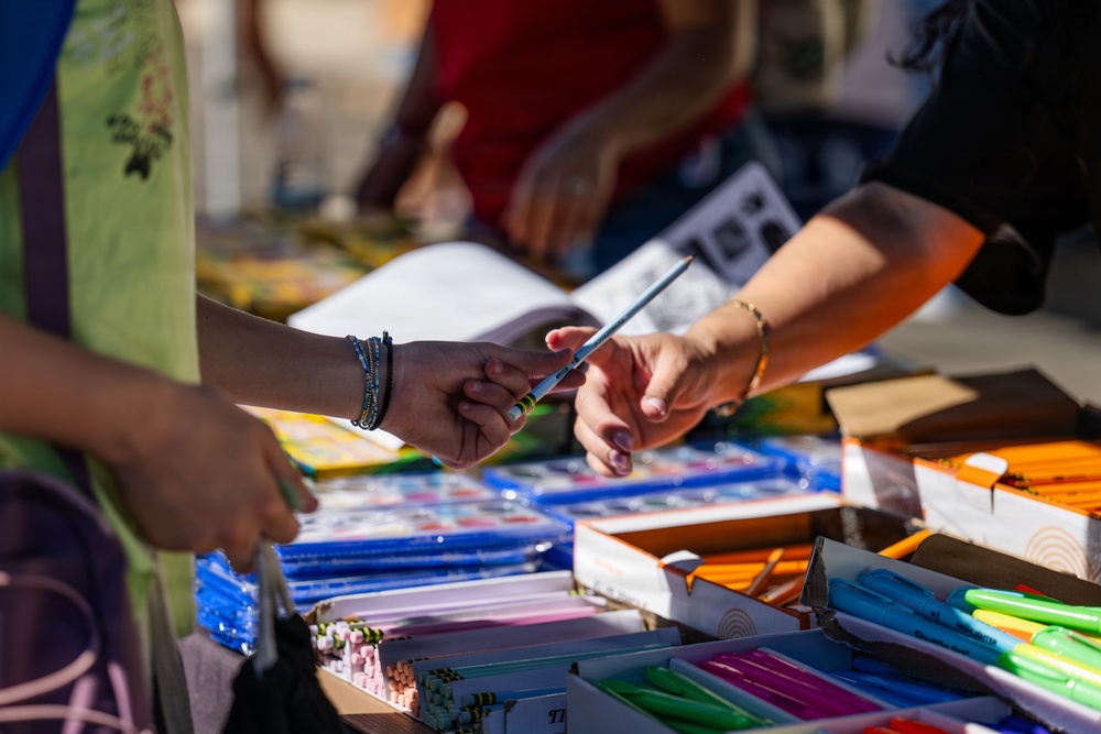 Two individuals are exchanging a pencil across a table covered with school supplies. The scene appears to be a school supply distribution event or donation drive, with multiple boxes of stationery items organized on the table. The foreground is in sharp focus while the background figures are blurred.