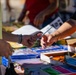 Two individuals are exchanging a pencil across a table covered with school supplies. The scene appears to be a school supply distribution event or donation drive, with multiple boxes of stationery items organized on the table. The foreground is in sharp focus while the background figures are blurred.