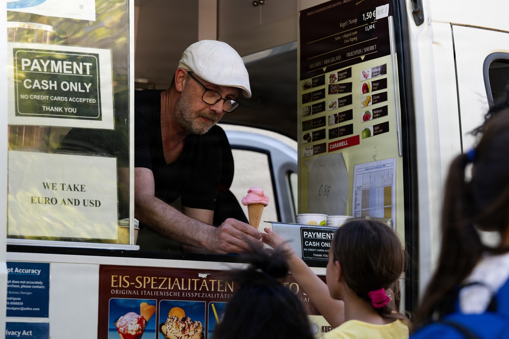 A middle-aged man wearing a white flat cap and glasses is serving a pink ice cream cone from an ice cream truck to a young girl. The truck displays signs indicating cash-only payment accepted in both Euro and USD, and features a menu board with various ice cream flavors. The signage on the truck includes German text reading 'Eis-Spezialitäten' (ice cream specialties) and 'Original Italienische Eisspezialitäten' (original Italian ice cream specialties).