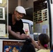 A middle-aged man wearing a white flat cap and glasses is serving a pink ice cream cone from an ice cream truck to a young girl. The truck displays signs indicating cash-only payment accepted in both Euro and USD, and features a menu board with various ice cream flavors. The signage on the truck includes German text reading 'Eis-Spezialitäten' (ice cream specialties) and 'Original Italienische Eisspezialitäten' (original Italian ice cream specialties).