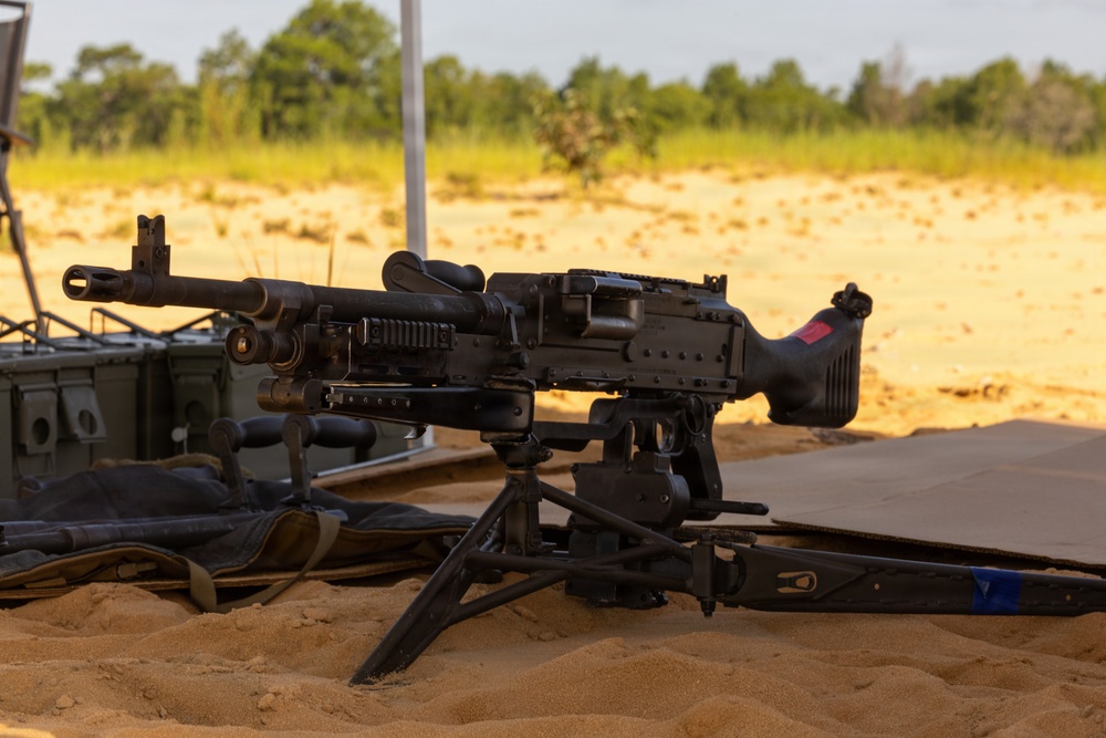 A general-purpose machine gun mounted on a bipod/tripod is positioned on sandy ground at what appears to be a military training range or firing position. The weapon is set up in a static firing configuration with supporting equipment visible nearby. The setting is an open sandy area with vegetation visible in the background under warm, golden-hour lighting.