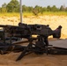 A general-purpose machine gun mounted on a bipod/tripod is positioned on sandy ground at what appears to be a military training range or firing position. The weapon is set up in a static firing configuration with supporting equipment visible nearby. The setting is an open sandy area with vegetation visible in the background under warm, golden-hour lighting.