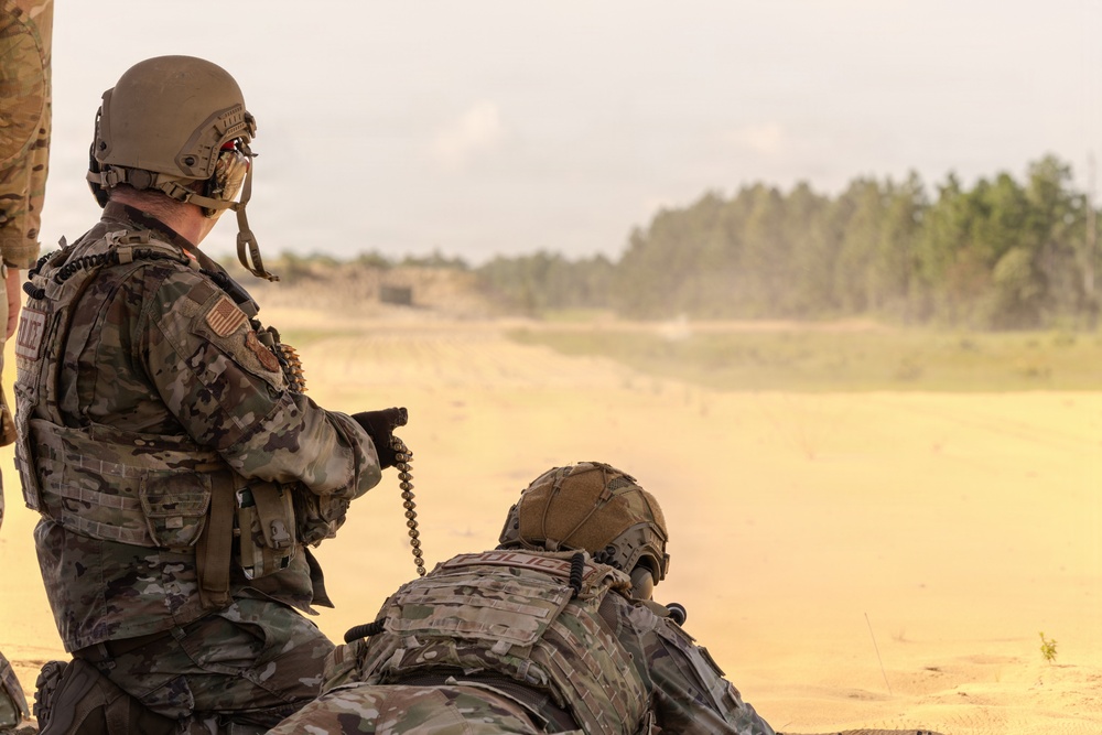 Two military personnel in full tactical gear are positioned on a sandy training range, with one kneeling and one in a prone position. Both are wearing multicam camouflage uniforms, tactical vests, and helmets, and appear to be observing or engaging a downrange area. The setting appears to be an outdoor live-fire or training range with a treeline visible in the background.