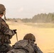 Two military personnel in full tactical gear are positioned on a sandy training range, with one kneeling and one in a prone position. Both are wearing multicam camouflage uniforms, tactical vests, and helmets, and appear to be observing or engaging a downrange area. The setting appears to be an outdoor live-fire or training range with a treeline visible in the background.