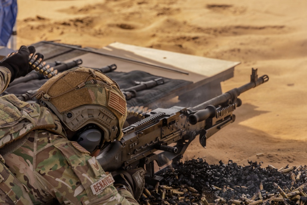 A U.S. military service member in full combat gear is firing a machine gun from a prone position in a desert environment. The soldier, identified by a name tape reading 'BENAVIDEZ,' is wearing MultiCam camouflage, a tactical helmet with a U.S. flag patch, and hearing protection. Spent brass casings and links are visible in the foreground, indicating active or recent firing.