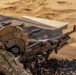 A U.S. military service member in full combat gear is firing a machine gun from a prone position in a desert environment. The soldier, identified by a name tape reading 'BENAVIDEZ,' is wearing MultiCam camouflage, a tactical helmet with a U.S. flag patch, and hearing protection. Spent brass casings and links are visible in the foreground, indicating active or recent firing.