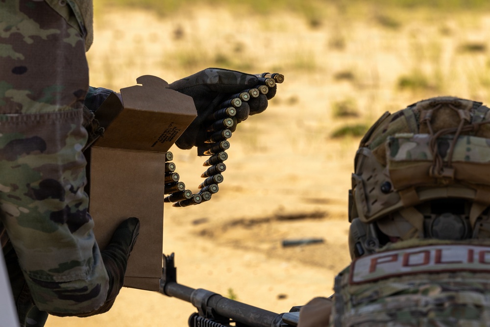 Two military personnel in MultiCam/OCP camouflage uniforms are engaged in a weapons training exercise at an outdoor range. One soldier is handling a linked ammunition belt from a cardboard ammunition box, while a second individual wearing a 'POLICE' patch on their helmet operates a crew-served weapon. The setting appears to be an arid, sandy training range during daylight hours.