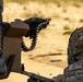 Two military personnel in MultiCam/OCP camouflage uniforms are engaged in a weapons training exercise at an outdoor range. One soldier is handling a linked ammunition belt from a cardboard ammunition box, while a second individual wearing a 'POLICE' patch on their helmet operates a crew-served weapon. The setting appears to be an arid, sandy training range during daylight hours.