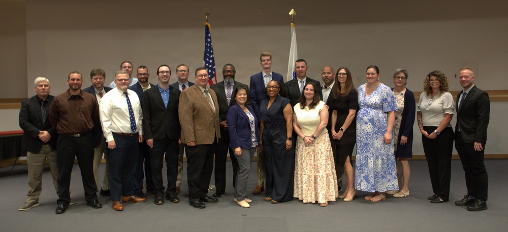 A large group of approximately 22 adults posed for a formal group photograph in what appears to be a government or institutional meeting room. Two flags are visible in the background — an American flag and what appears to be a state flag. The group is dressed in business and business-casual attire, suggesting a professional event such as a graduation, swearing-in ceremony, or organizational meeting.
