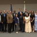 A large group of approximately 22 adults posed for a formal group photograph in what appears to be a government or institutional meeting room. Two flags are visible in the background — an American flag and what appears to be a state flag. The group is dressed in business and business-casual attire, suggesting a professional event such as a graduation, swearing-in ceremony, or organizational meeting.