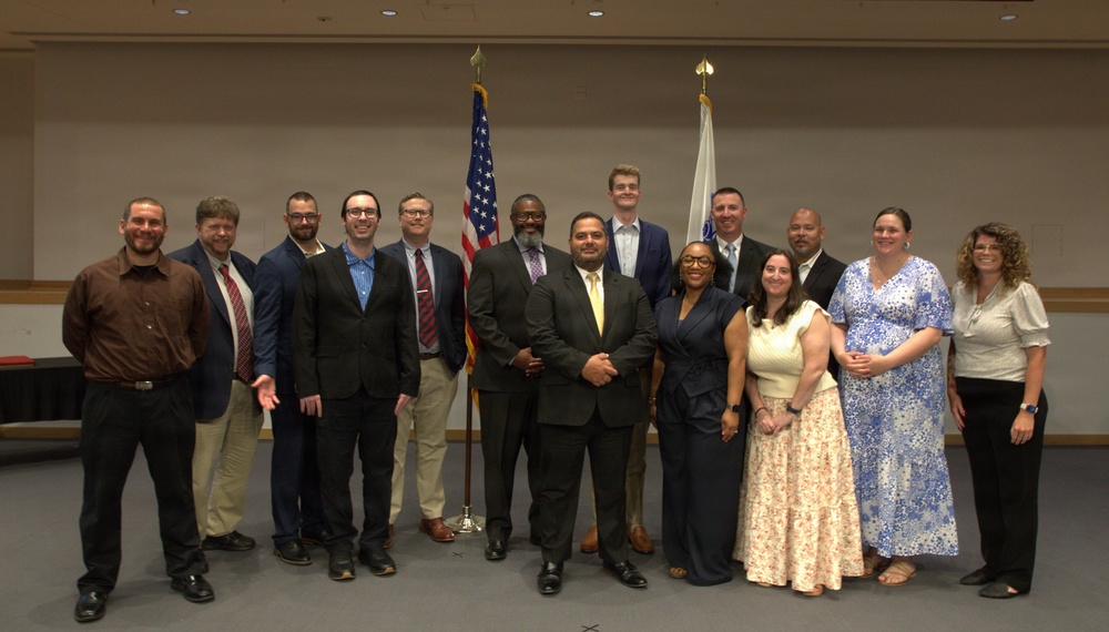 A group of approximately 14 adults posed for a formal group photograph in what appears to be a conference or civic hall setting. The individuals are dressed in business and business-casual attire, standing in two rows in front of an American flag and what appears to be a state flag. The setting and attire suggest an official ceremony, graduation, or professional recognition event.