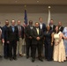 A group of approximately 14 adults posed for a formal group photograph in what appears to be a conference or civic hall setting. The individuals are dressed in business and business-casual attire, standing in two rows in front of an American flag and what appears to be a state flag. The setting and attire suggest an official ceremony, graduation, or professional recognition event.