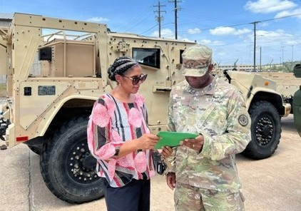 A U.S. Army soldier in OCP (Operational Camouflage Pattern) uniform is reviewing a green document or folder with a civilian woman in a colorful pink and white blouse. They are standing in front of a tan military HMMWV (Humvee) in what appears to be a military installation or motor pool area. The setting suggests an administrative or inspection-related interaction outdoors.