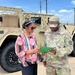 A U.S. Army soldier in OCP (Operational Camouflage Pattern) uniform is reviewing a green document or folder with a civilian woman in a colorful pink and white blouse. They are standing in front of a tan military HMMWV (Humvee) in what appears to be a military installation or motor pool area. The setting suggests an administrative or inspection-related interaction outdoors.