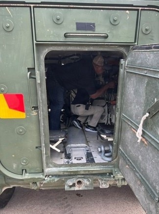 The image shows the interior of a military-green vehicle, viewed through an open rear door. A person wearing a hat and light-colored trousers is seated or crouching inside the cramped compartment. The vehicle appears to be a military or utility transport, with metal flooring, tie-down hardware, and a red-and-yellow hazard marking visible on the exterior.
