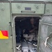 The image shows the interior of a military-green vehicle, viewed through an open rear door. A person wearing a hat and light-colored trousers is seated or crouching inside the cramped compartment. The vehicle appears to be a military or utility transport, with metal flooring, tie-down hardware, and a red-and-yellow hazard marking visible on the exterior.