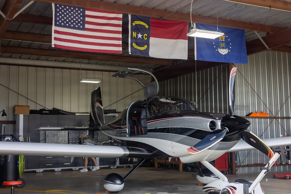 A sleek, modern light aircraft with a dark color scheme and gull-wing doors open is parked inside a metal hangar. Three flags are displayed prominently on the back wall: the United States flag, the North Carolina state flag, and the United States Air Force flag. A person is partially visible near the rear of the aircraft, and the hangar contains tool storage cabinets and equipment.