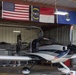A sleek, modern light aircraft with a dark color scheme and gull-wing doors open is parked inside a metal hangar. Three flags are displayed prominently on the back wall: the United States flag, the North Carolina state flag, and the United States Air Force flag. A person is partially visible near the rear of the aircraft, and the hangar contains tool storage cabinets and equipment.