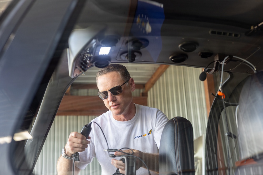 A middle-aged man wearing sunglasses and a white 'Atlas Air' branded t-shirt is working inside or near the cockpit of a small aircraft. He is holding what appears to be an inspection tool or borescope with a cable attached, suggesting he is performing a maintenance or inspection task. The photo is taken from outside the aircraft looking through the windscreen/canopy.