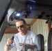 A middle-aged man wearing sunglasses and a white 'Atlas Air' branded t-shirt is working inside or near the cockpit of a small aircraft. He is holding what appears to be an inspection tool or borescope with a cable attached, suggesting he is performing a maintenance or inspection task. The photo is taken from outside the aircraft looking through the windscreen/canopy.