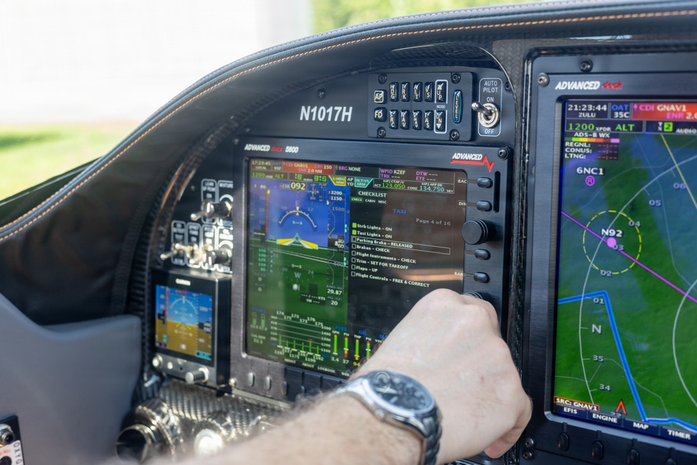 The image shows the cockpit interior of a small aircraft with tail number N1017H, featuring Advanced Cock 8800 avionics displays. A pilot's hand wearing a watch is interacting with the central EFIS/MFD screen, which is displaying a TAXI checklist on page 4 of 16. The cockpit is equipped with modern glass panel avionics including navigation displays, autopilot controls, and a moving map.