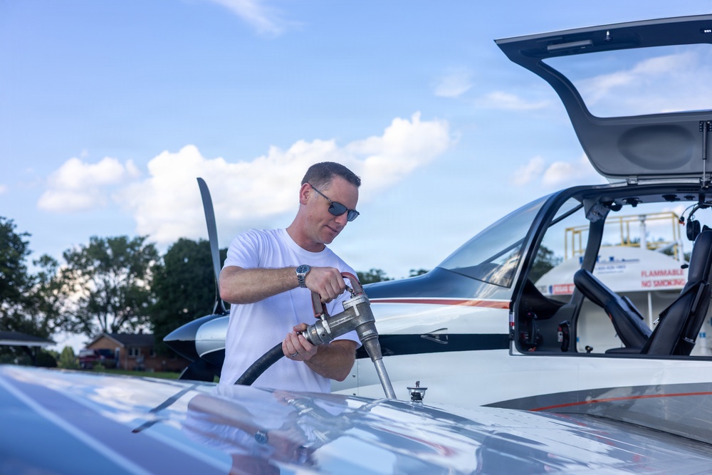 A man in a white t-shirt and sunglasses is fueling a small private aircraft on what appears to be a general aviation airport ramp. He is handling a fuel nozzle inserted into the wing fuel port of a modern, sleek single-engine propeller aircraft. The canopy/door of the aircraft is open, and a fuel truck or fuel station is visible in the background.