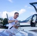 A man in a white t-shirt and sunglasses is fueling a small private aircraft on what appears to be a general aviation airport ramp. He is handling a fuel nozzle inserted into the wing fuel port of a modern, sleek single-engine propeller aircraft. The canopy/door of the aircraft is open, and a fuel truck or fuel station is visible in the background.