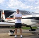 A man stands confidently in front of a modern single-engine propeller aircraft on an airport tarmac. He is posing with arms crossed, smiling, in front of what appears to be a sleek, white and orange private plane with registration number N1917U. The setting appears to be a small general aviation airport with a hangar visible in the background.
