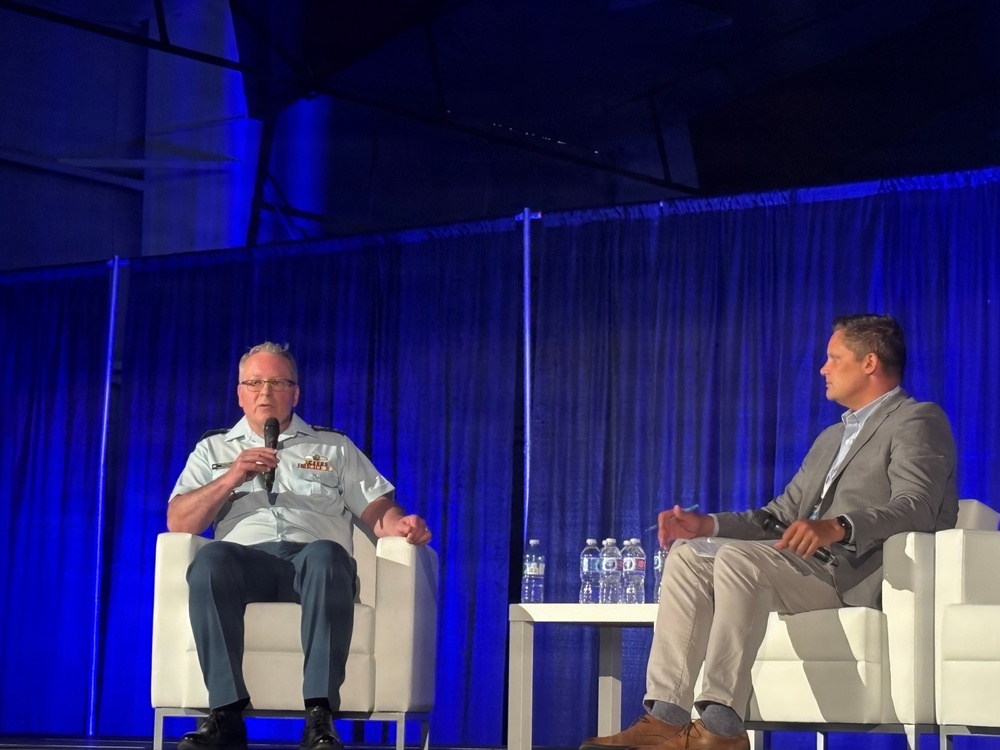 Two men are seated on a stage in white chairs during what appears to be a professional conference or panel discussion. One man is wearing a military uniform and speaking into a handheld microphone, while the other is dressed in civilian business casual attire and listening. The stage is lit with blue lighting and dark curtain backdrop.