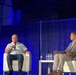 Two men are seated on a stage in white chairs during what appears to be a professional conference or panel discussion. One man is wearing a military uniform and speaking into a handheld microphone, while the other is dressed in civilian business casual attire and listening. The stage is lit with blue lighting and dark curtain backdrop.