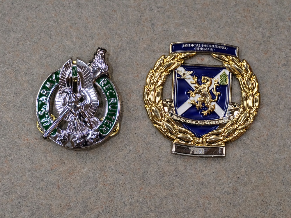 The image shows two U.S. Army military badges/insignia placed on a gray surface. The left badge is a silver and green U.S. Army Recruiter badge, while the right is a gold and blue Medical Recruiting Brigade badge with the number 763. Both badges are in good condition and clearly visible.