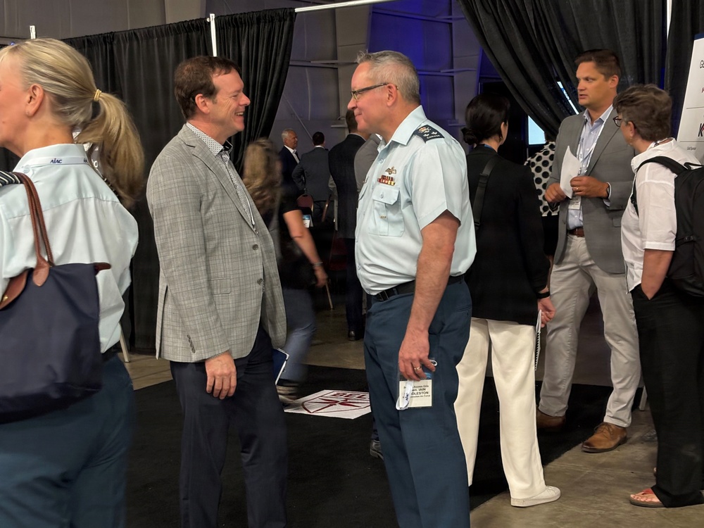 The image shows a professional conference or expo setting where attendees are networking. A military officer in a light blue uniform is engaged in conversation with a civilian in a gray blazer. The event appears to be a defense or aerospace industry gathering based on the military presence and conference environment.