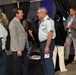 The image shows a professional conference or expo setting where attendees are networking. A military officer in a light blue uniform is engaged in conversation with a civilian in a gray blazer. The event appears to be a defense or aerospace industry gathering based on the military presence and conference environment.