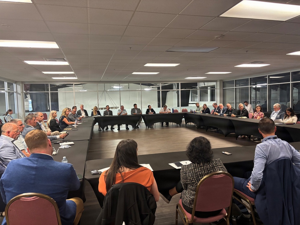 A large formal meeting or roundtable discussion is taking place in a modern conference room with floor-to-ceiling windows. Approximately 25-35 participants are seated around a large U-shaped or rectangular table arrangement, suggesting a business, government, or organizational gathering. The setting appears professional with overhead fluorescent lighting and a darkened exterior visible through the windows.