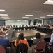 A large formal meeting or roundtable discussion is taking place in a modern conference room with floor-to-ceiling windows. Approximately 25-35 participants are seated around a large U-shaped or rectangular table arrangement, suggesting a business, government, or organizational gathering. The setting appears professional with overhead fluorescent lighting and a darkened exterior visible through the windows.