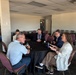 Five individuals are seated around a round table with a black tablecloth in what appears to be a conference or event venue room. They appear to be engaged in a small group discussion or meeting, with one woman gesturing as she speaks. The room contains multiple additional round tables with empty chairs, suggesting a larger event space.