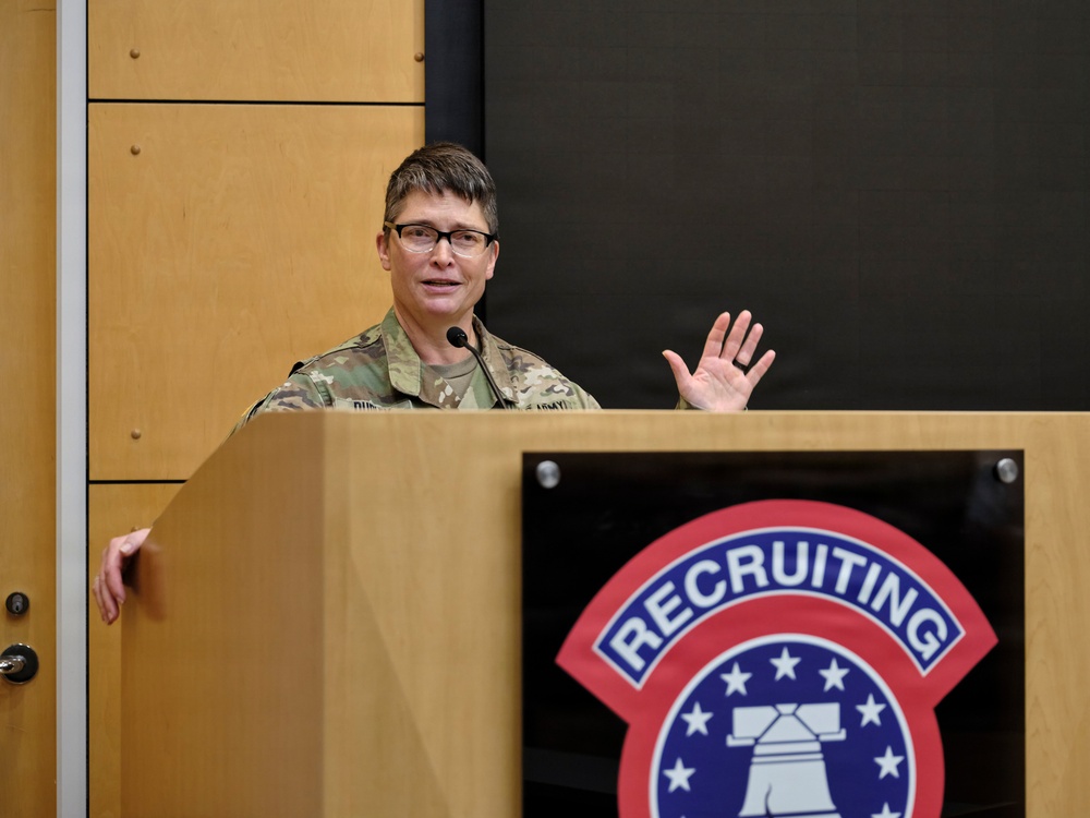 A U.S. Army officer in camouflage uniform is speaking at a podium during what appears to be an official military briefing or presentation. The podium displays the U.S. Army Recruiting Command insignia. The speaker is gesturing with one raised hand while addressing an audience.