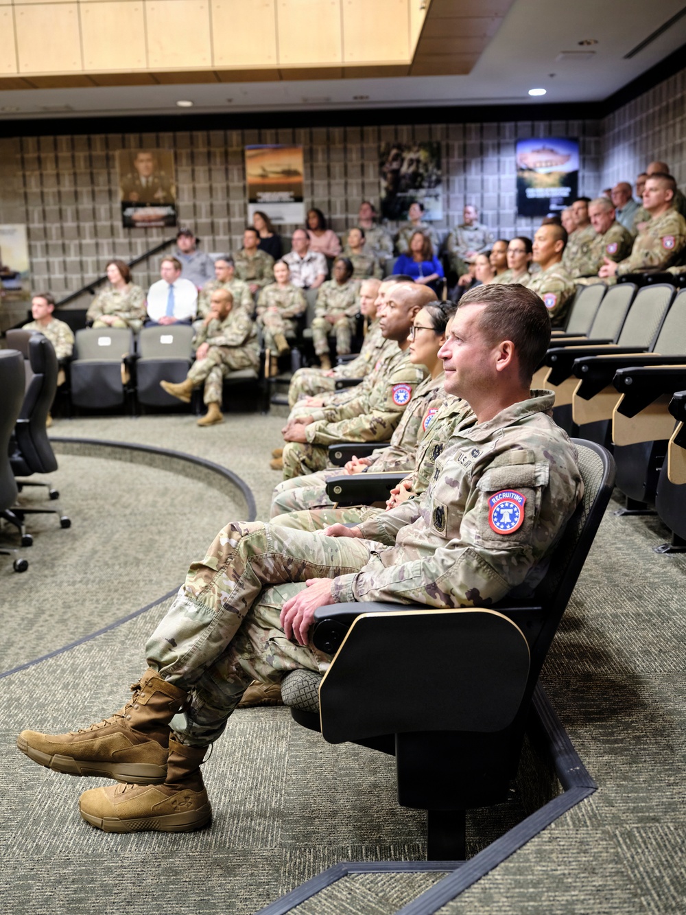 A group of U.S. Army personnel and some civilians are seated in a tiered auditorium or briefing room, appearing to attend a presentation or briefing. The majority of attendees are wearing Operational Camouflage Pattern (OCP) uniforms, with at least one civilian in business attire visible. The foreground soldier prominently displays a 'Recruiting' shoulder patch consistent with U.S. Army Recruiting Command.