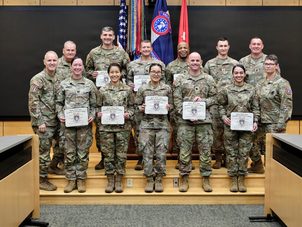 A group of U.S. Army soldiers pose for a formal group photograph in what appears to be a military briefing or classroom facility. Several soldiers in the front row are holding certificates from what appears to be the United States Army Recruiting and Retention College. The image captures what appears to be a graduation or recognition ceremony.