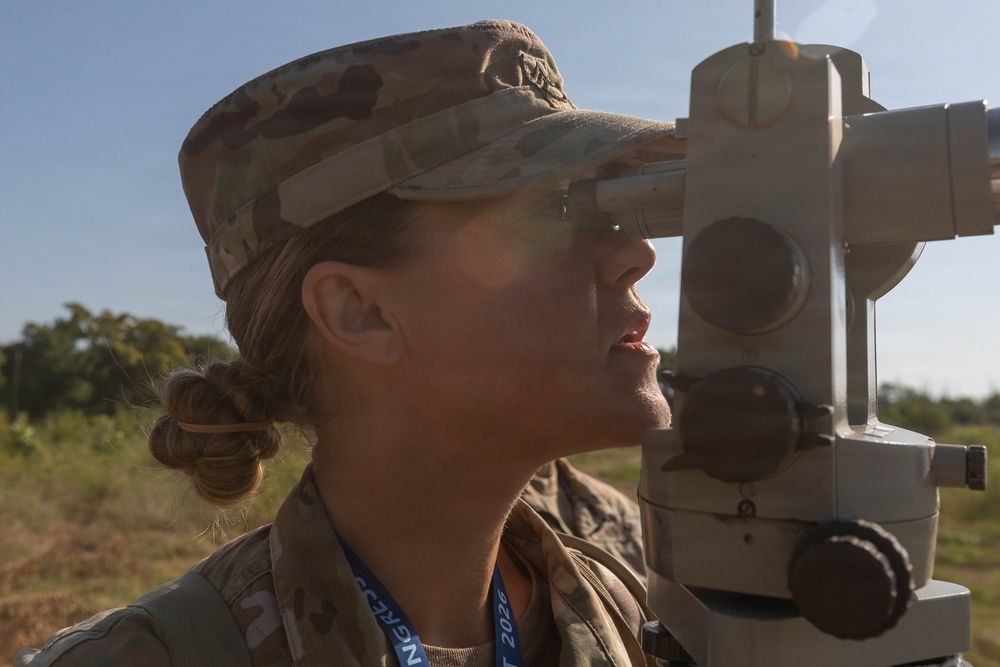 A female military service member in MultiCam/OCP camouflage uniform is looking through the eyepiece of a surveying or optical instrument, likely a theodolite or aiming circle, in an outdoor field setting. She is wearing a patrol cap and has a blue lanyard/badge around her neck with visible text including '2026'. The image is a close-up profile shot taken in bright natural sunlight.
