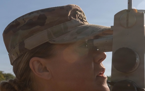 A female military service member in MultiCam/OCP camouflage uniform is looking through the eyepiece of a surveying or optical instrument, likely a theodolite or aiming circle, in an outdoor field setting. She is wearing a patrol cap and has a blue lanyard/badge around her neck with visible text including '2026'. The image is a close-up profile shot taken in bright natural sunlight.