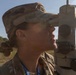 A female military service member in MultiCam/OCP camouflage uniform is looking through the eyepiece of a surveying or optical instrument, likely a theodolite or aiming circle, in an outdoor field setting. She is wearing a patrol cap and has a blue lanyard/badge around her neck with visible text including '2026'. The image is a close-up profile shot taken in bright natural sunlight.