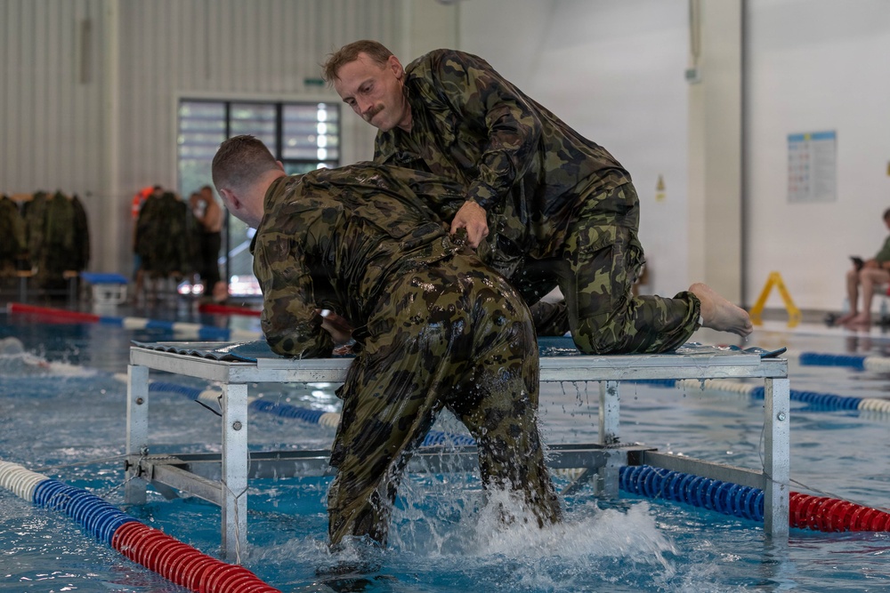 Two military personnel in wet camouflage uniforms are engaged in a water survival or combat training exercise at an indoor swimming pool facility. One individual appears to be assisting or restraining the other on a metal platform positioned over the pool. Additional uniformed personnel are visible in the background observing or awaiting their turn.