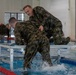 Two military personnel in wet camouflage uniforms are engaged in a water survival or combat training exercise at an indoor swimming pool facility. One individual appears to be assisting or restraining the other on a metal platform positioned over the pool. Additional uniformed personnel are visible in the background observing or awaiting their turn.