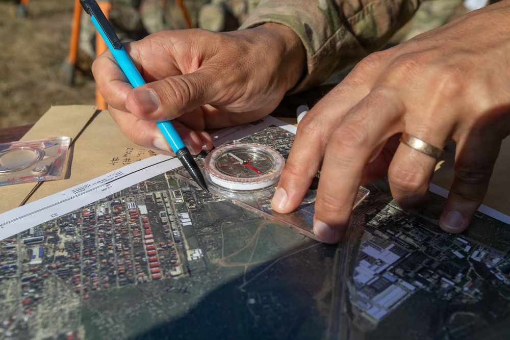 A person wearing military camouflage uniform is using a baseplate compass and a blue pencil to plot or mark positions on an aerial/satellite map. The individual is wearing a gold wedding band on their right hand. The activity appears to be military land navigation or operational planning conducted outdoors.