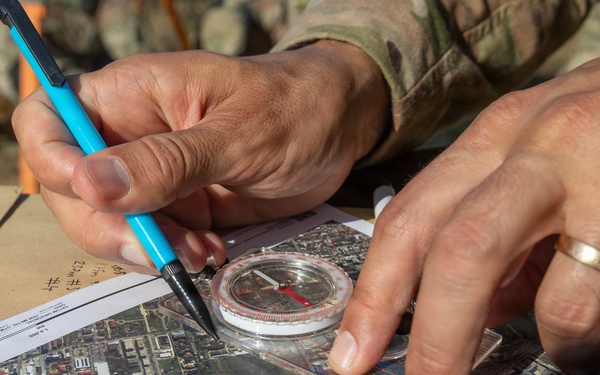 A person wearing military camouflage uniform is using a baseplate compass and a blue pencil to plot or mark positions on an aerial/satellite map. The individual is wearing a gold wedding band on their right hand. The activity appears to be military land navigation or operational planning conducted outdoors.