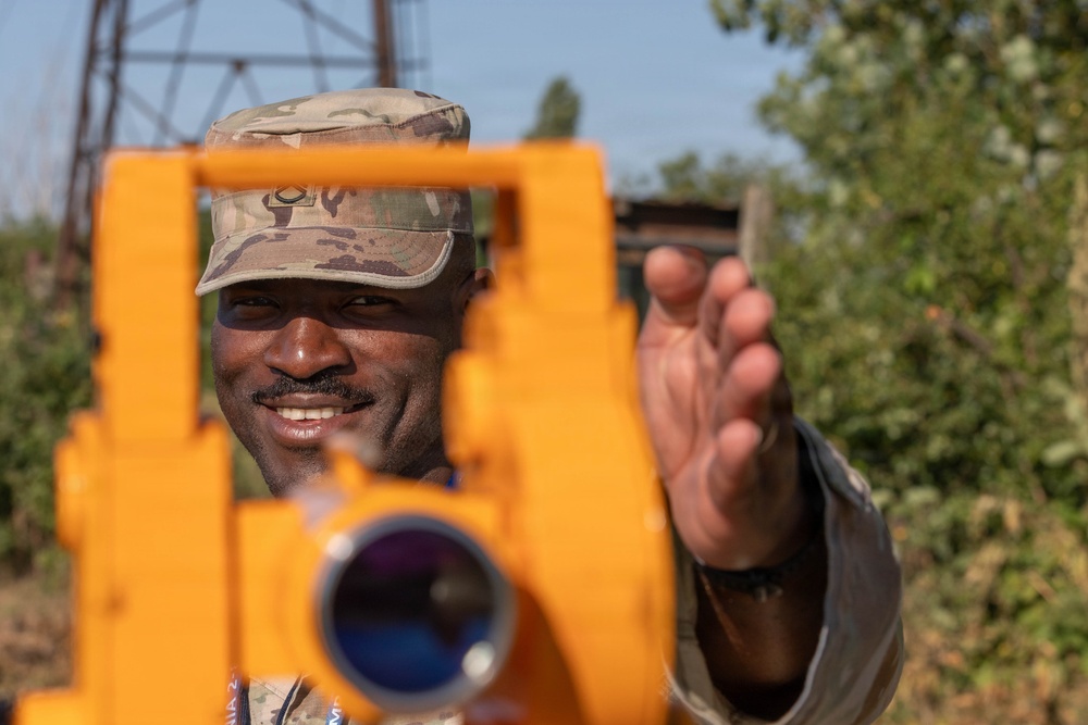A military service member in camouflage uniform and patrol cap is smiling while operating or adjusting a bright orange surveying instrument, likely a theodolite or transit level. The soldier appears to be conducting land surveying or engineering work in an outdoor environment. The image is taken from the perspective of looking through or past the instrument toward the operator.