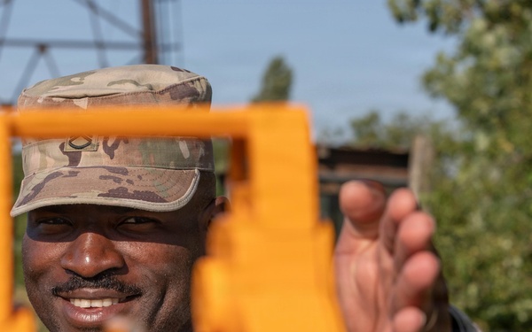 A military service member in camouflage uniform and patrol cap is smiling while operating or adjusting a bright orange surveying instrument, likely a theodolite or transit level. The soldier appears to be conducting land surveying or engineering work in an outdoor environment. The image is taken from the perspective of looking through or past the instrument toward the operator.